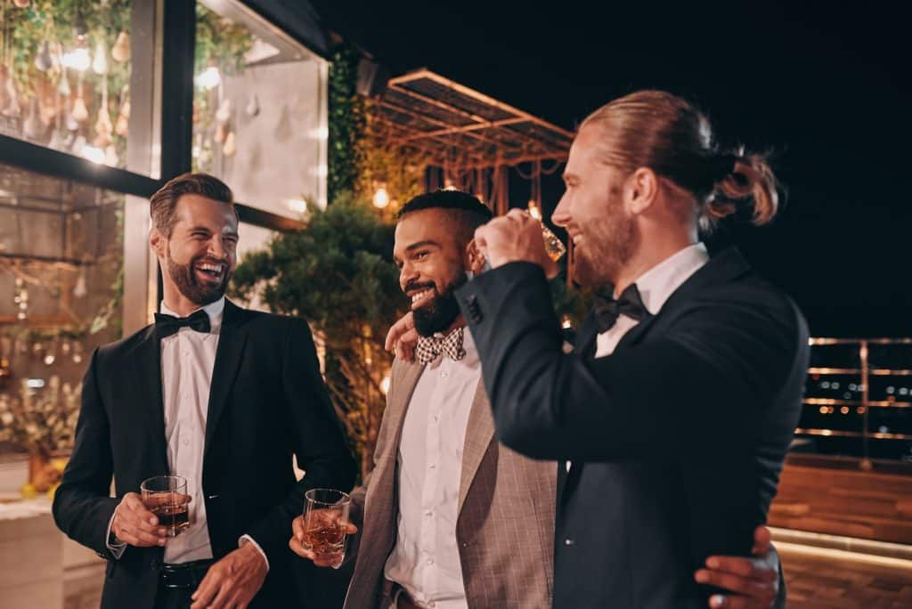 Three happy men in suits holding glasses with whiskey while spending time on party
