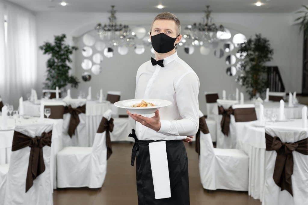 Young happy waiter wearing protective face mask while serving food in a restaurant